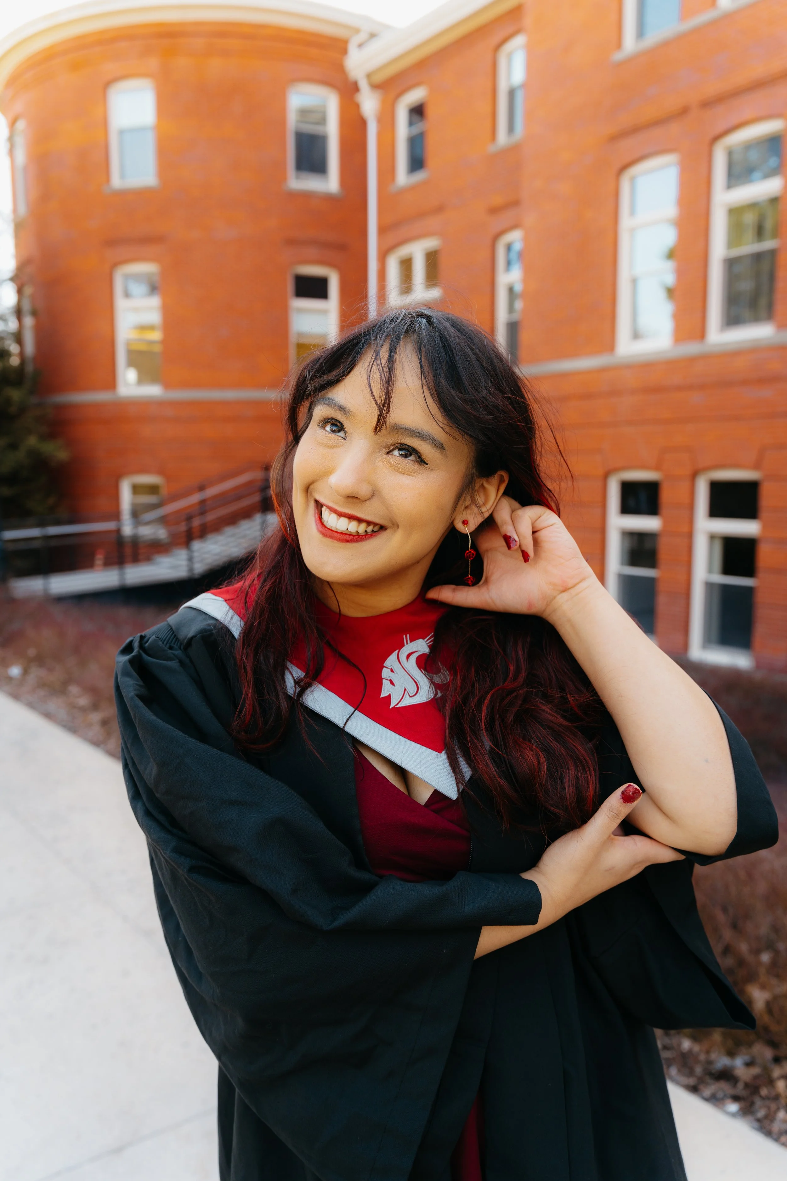 A smiling woman dressed in a graduation gown and stole, outdoors near a red brick building with multiple windows, during daytime.
