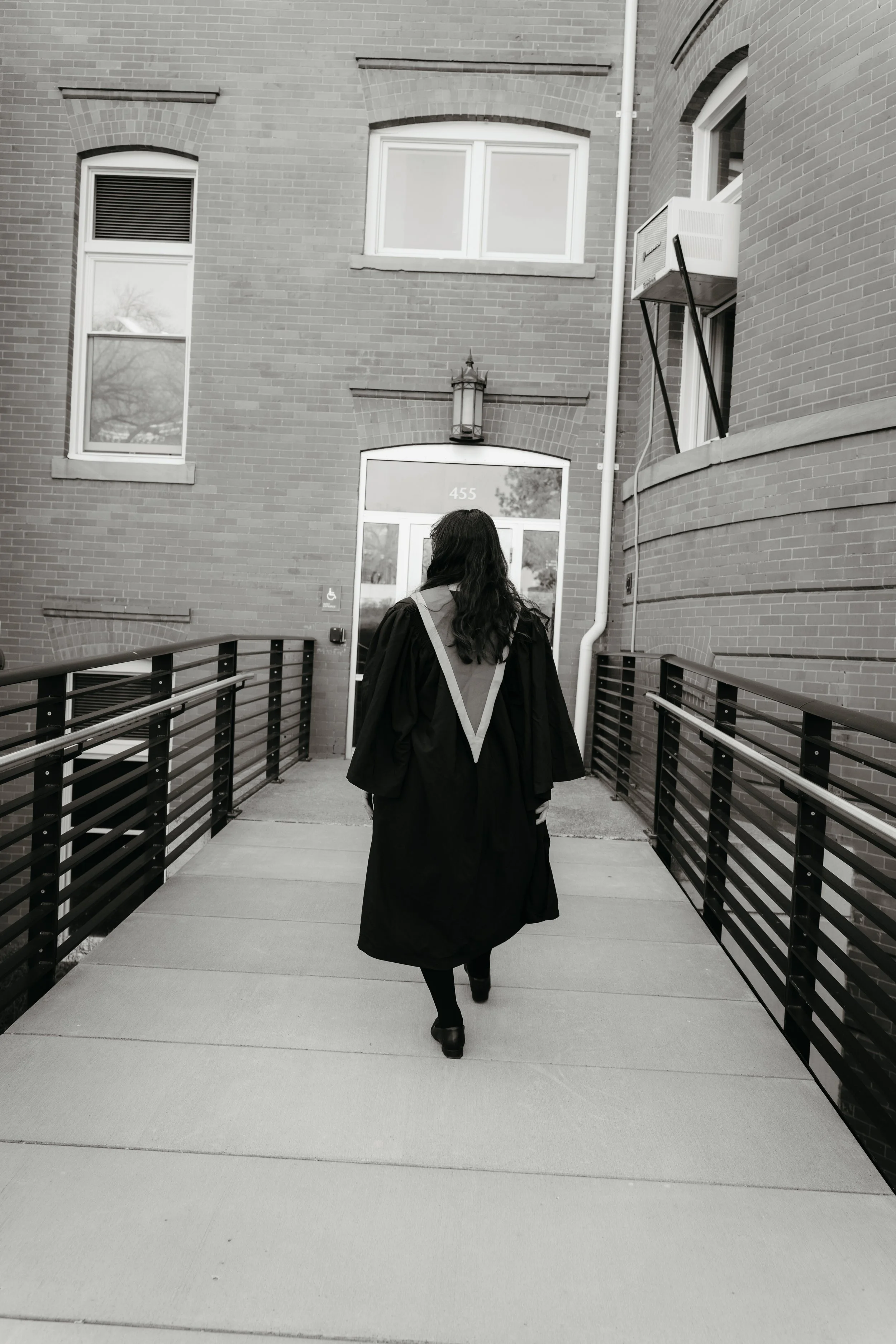 A person dressed in a graduation gown walking away towards a brick building with the number 455 on the door.