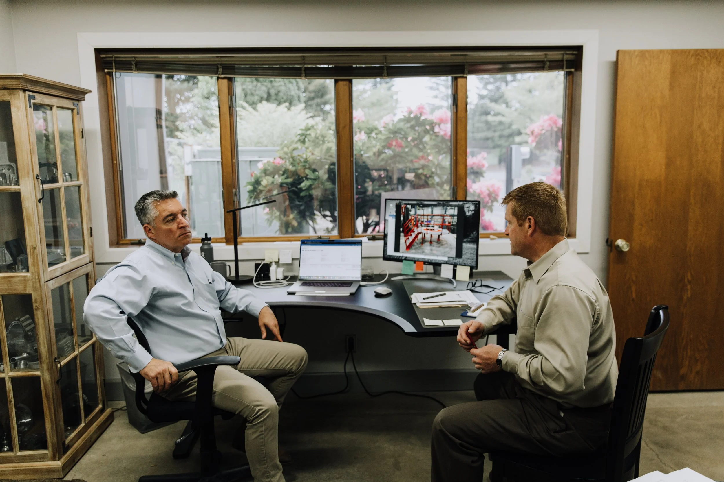 Two men in business attire having a discussion in an office with a view of trees and pink flowers outside the window. There are two computer monitors, scattered papers, and a wooden cabinet in the room.