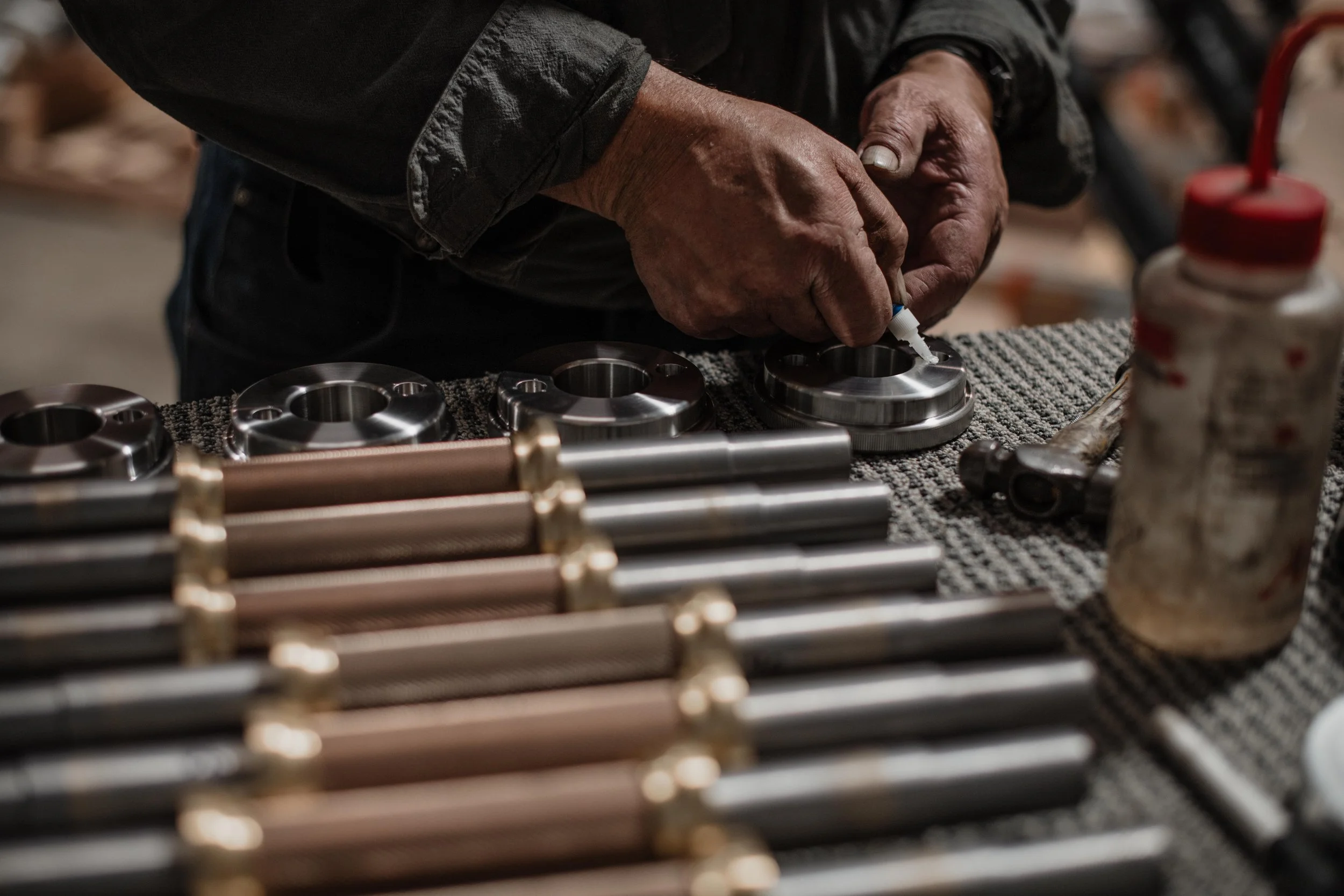 A person working with metal parts, applying lubricant or glue to circular metal components on a textured work surface surrounded by various metal rods and tools.