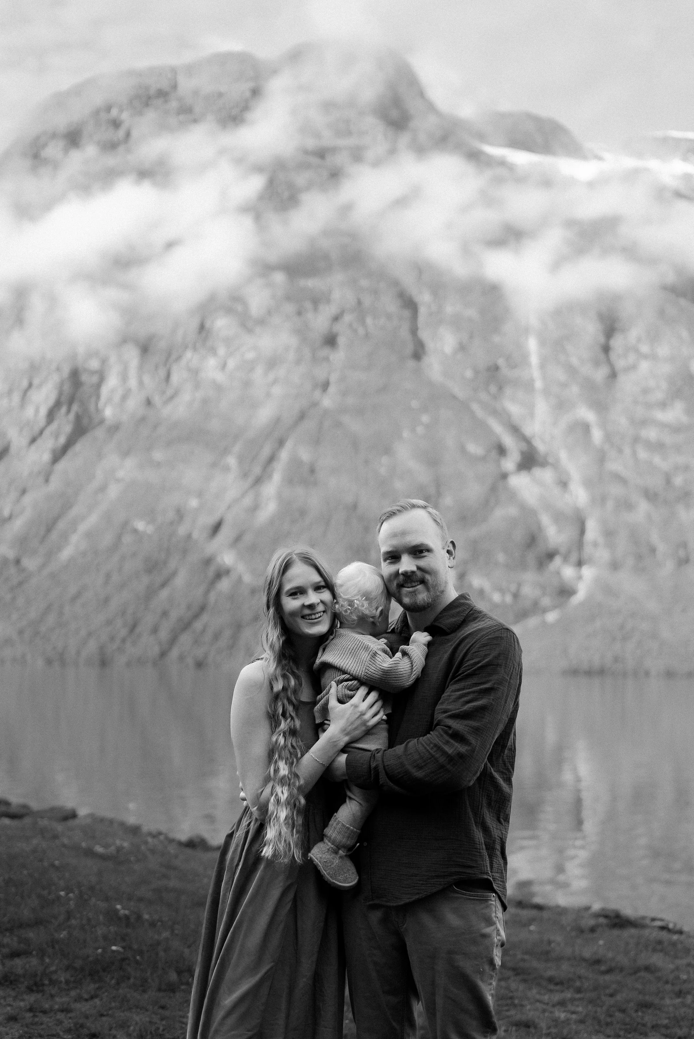 A family of three, including a woman, a man, and a child, poses outdoors near a body of water with a mountain landscape in the background.