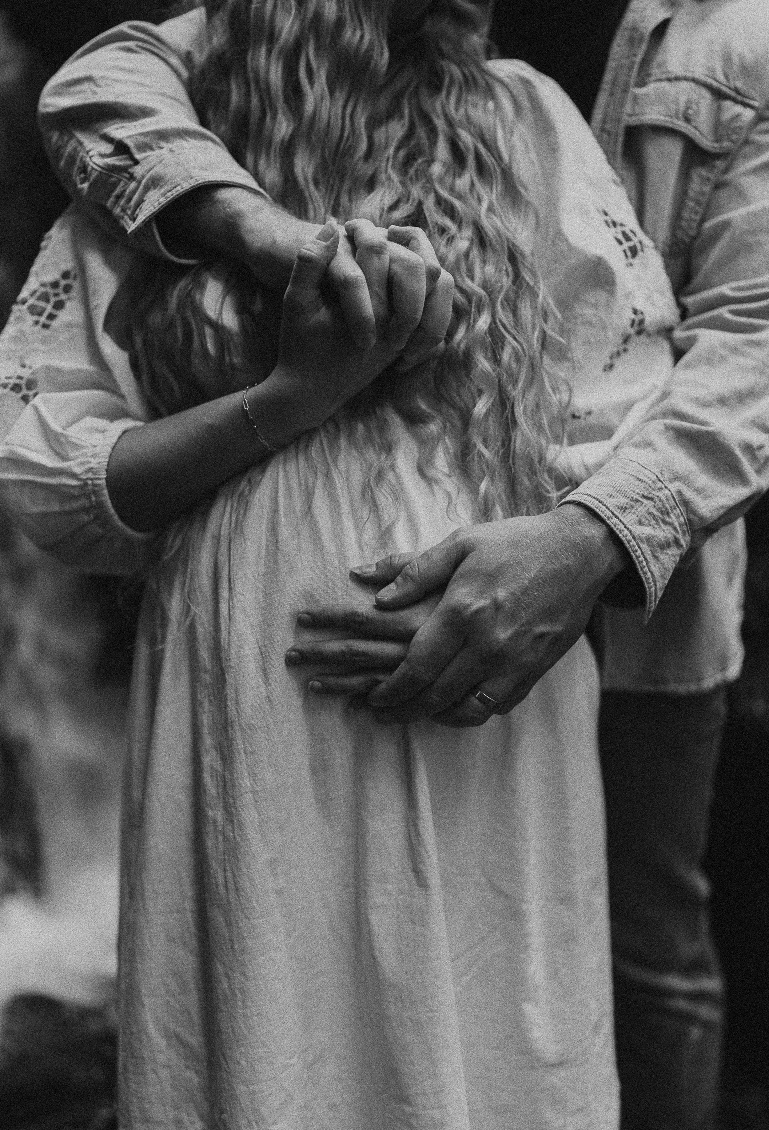 A black and white photo of a couple holding hands, with the woman's hand resting on her pregnant belly and the man's hand gently placed on top, both wearing rings.
