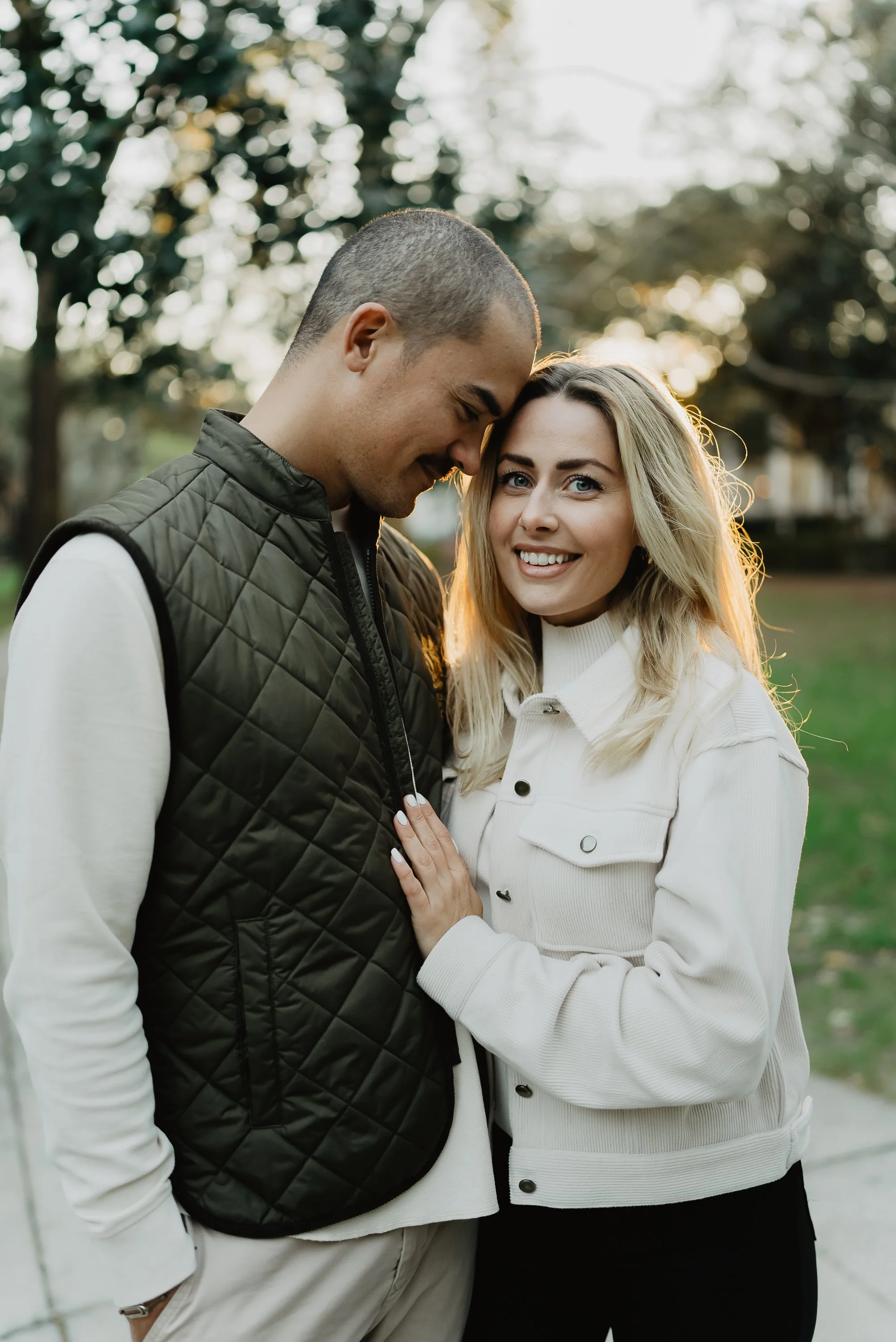 A smiling woman and a man are standing close together outdoors, with the woman touching the man's chest. They are in a park with trees and sunlight in the background.