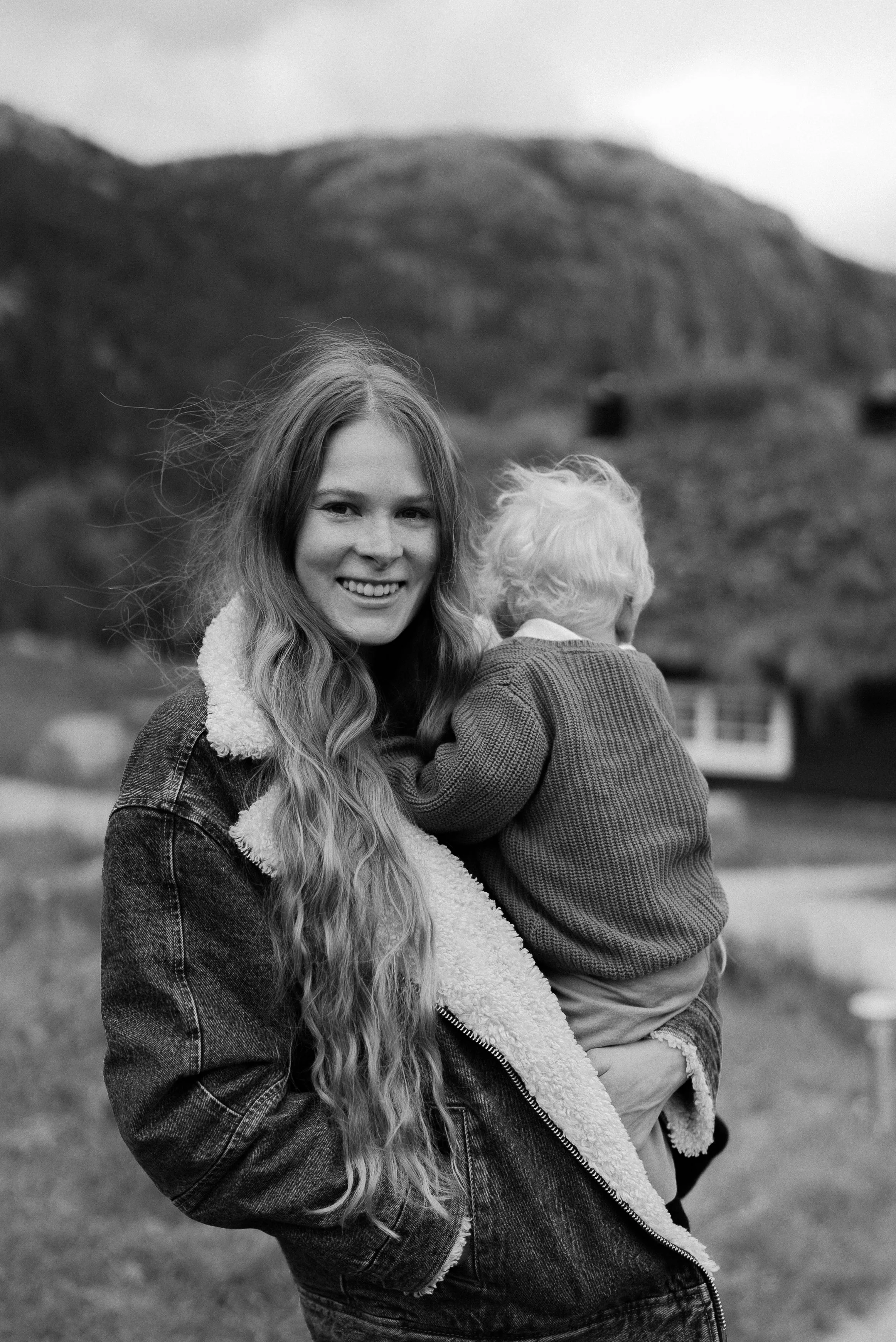 A woman with long wavy hair smiling while holding a young child in her arms outdoors with mountains in the background.