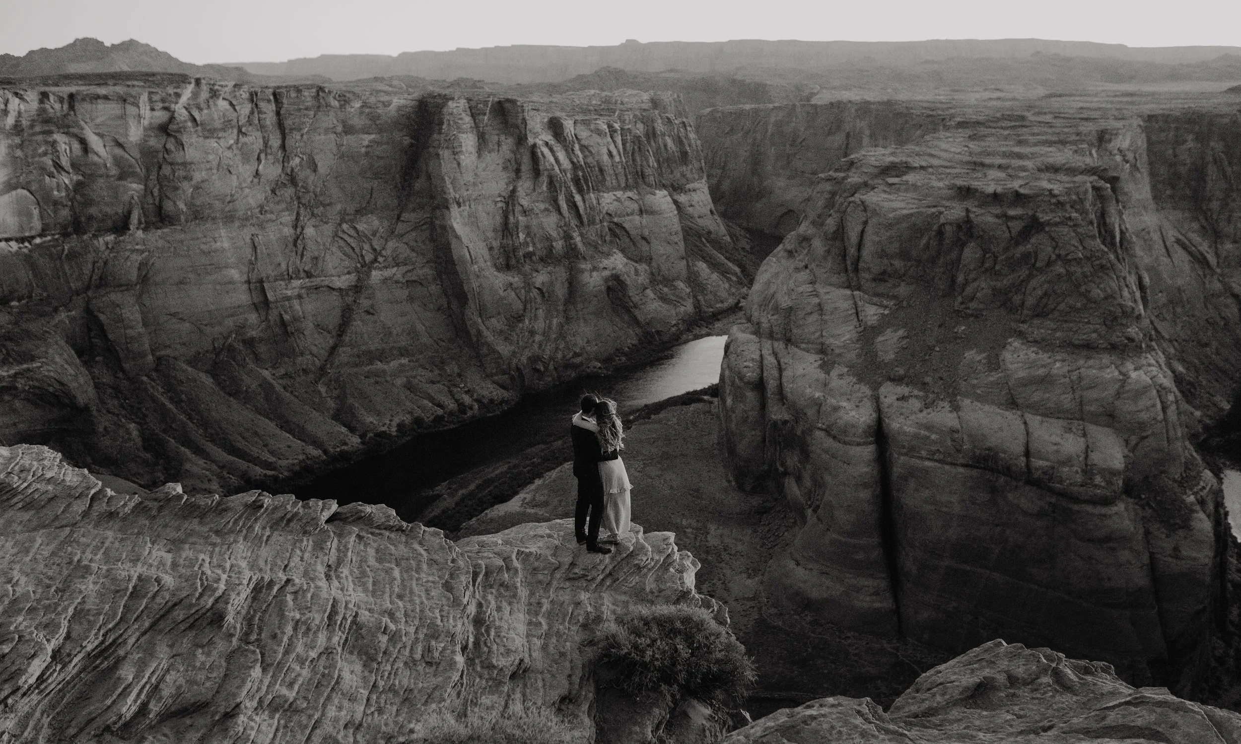 A couple embracing at the edge of a cliff overlooking a canyon with a river, in black and white.