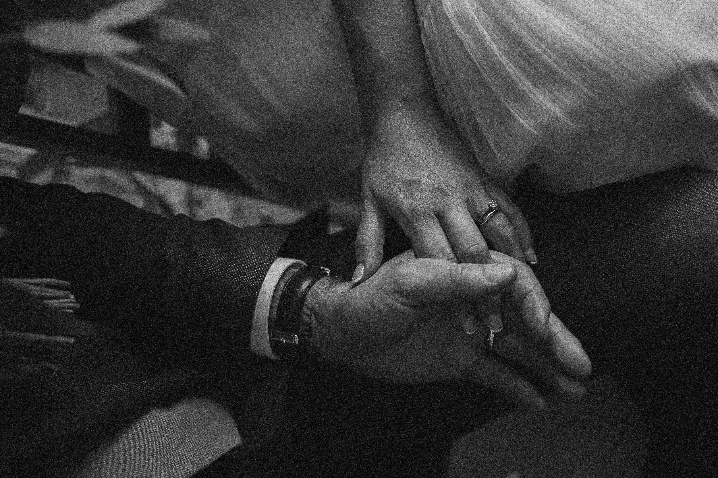 Close-up of two people holding hands, one wearing a dark suit and the other wearing a striped skirt, with wedding rings visible on their fingers, suggesting a romantic or wedding context.