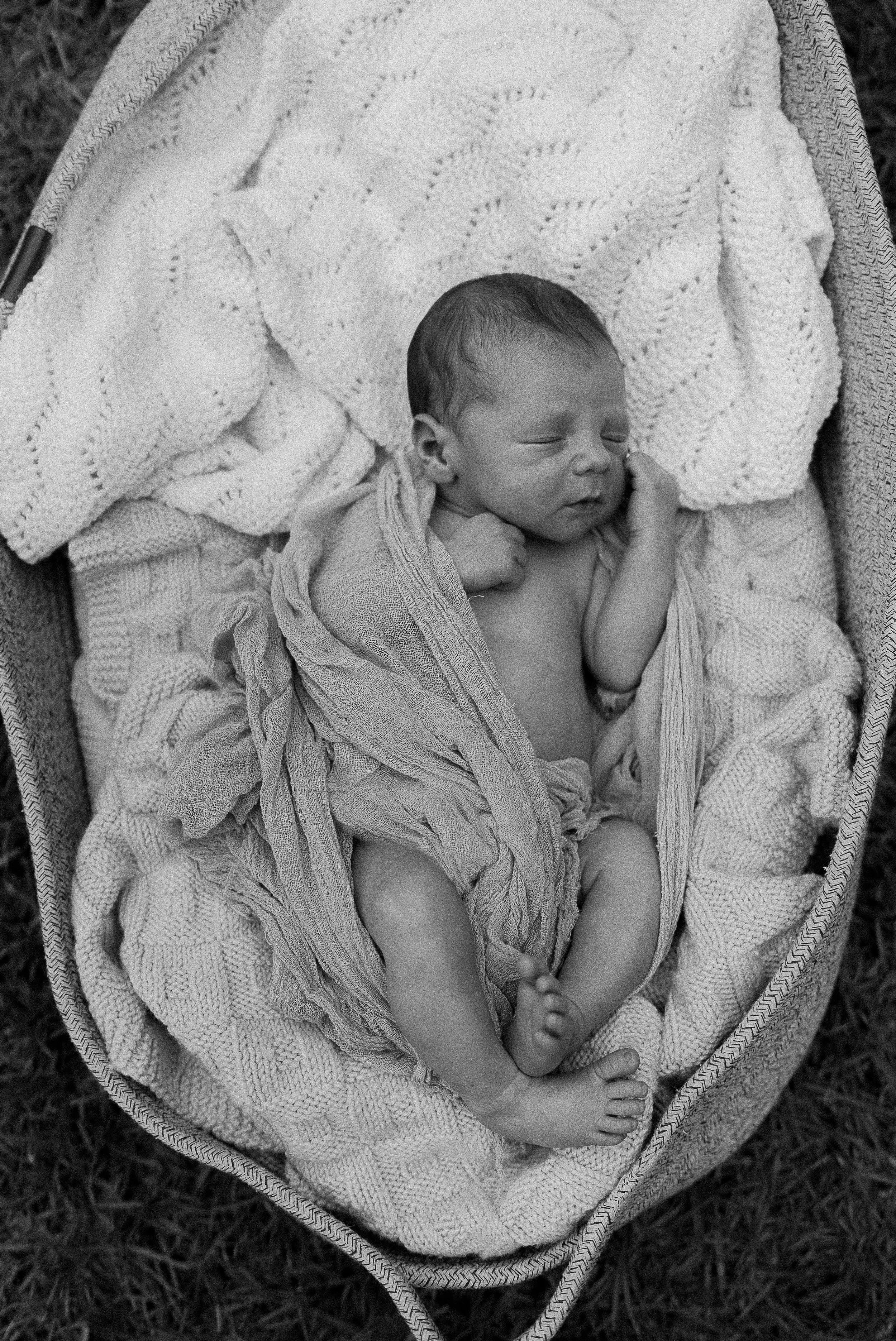 A newborn baby sleeping in a bassinet, wrapped in a soft blanket, with a knitted blanket behind their head.