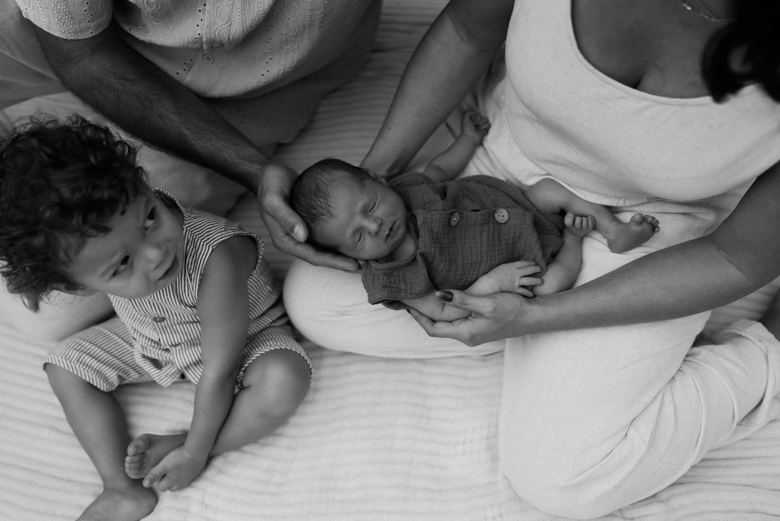 A woman holding a newborn baby and a young child sitting on a bed, all in black and white.