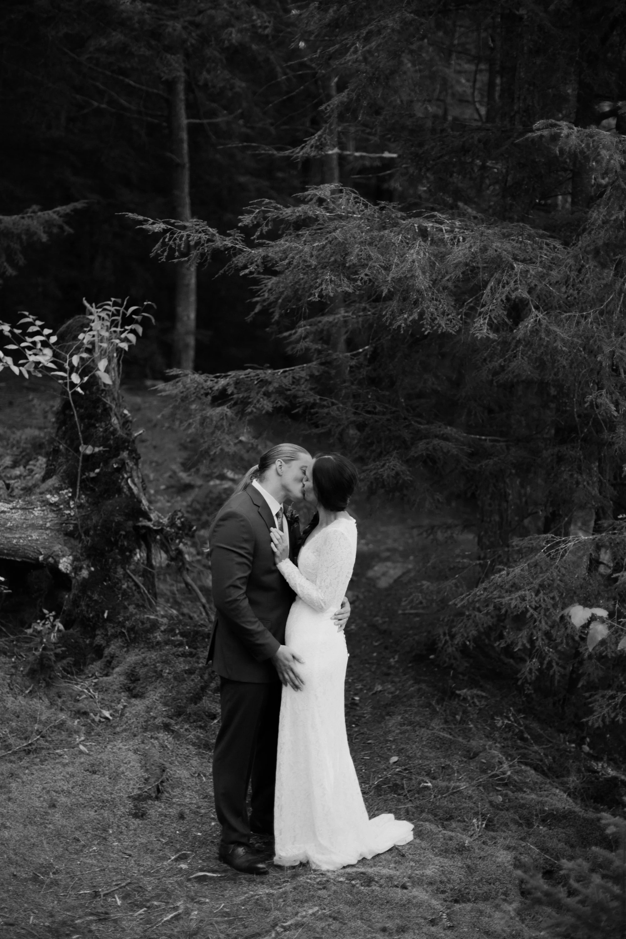 A black and white photograph of a couple in wedding attire sharing a kiss in a forest, surrounded by trees and natural scenery.