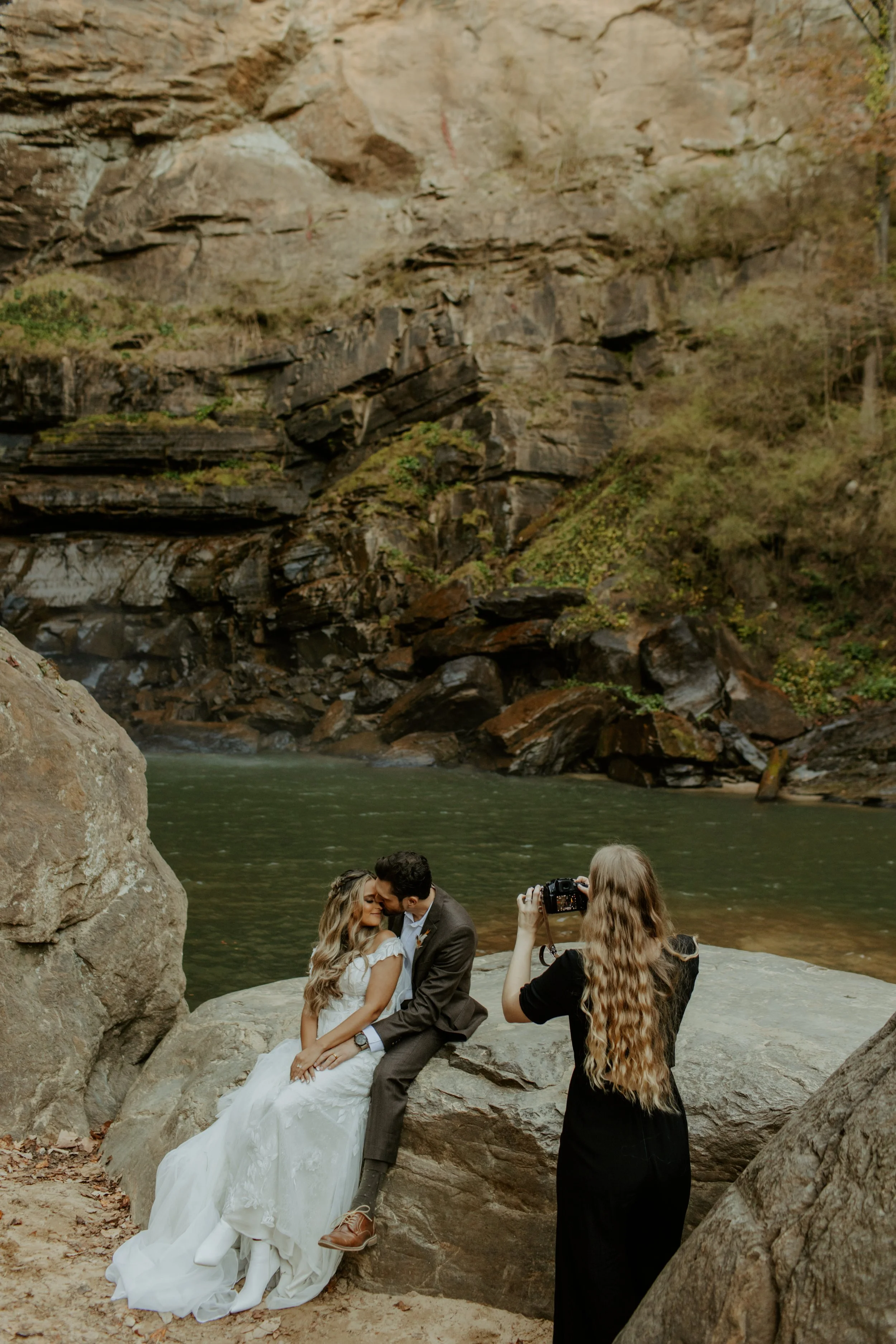 Couple dressed in wedding attire sitting on a large rock by a river, with a woman taking their photograph in a forested area with rocks and a waterfall in the background.
