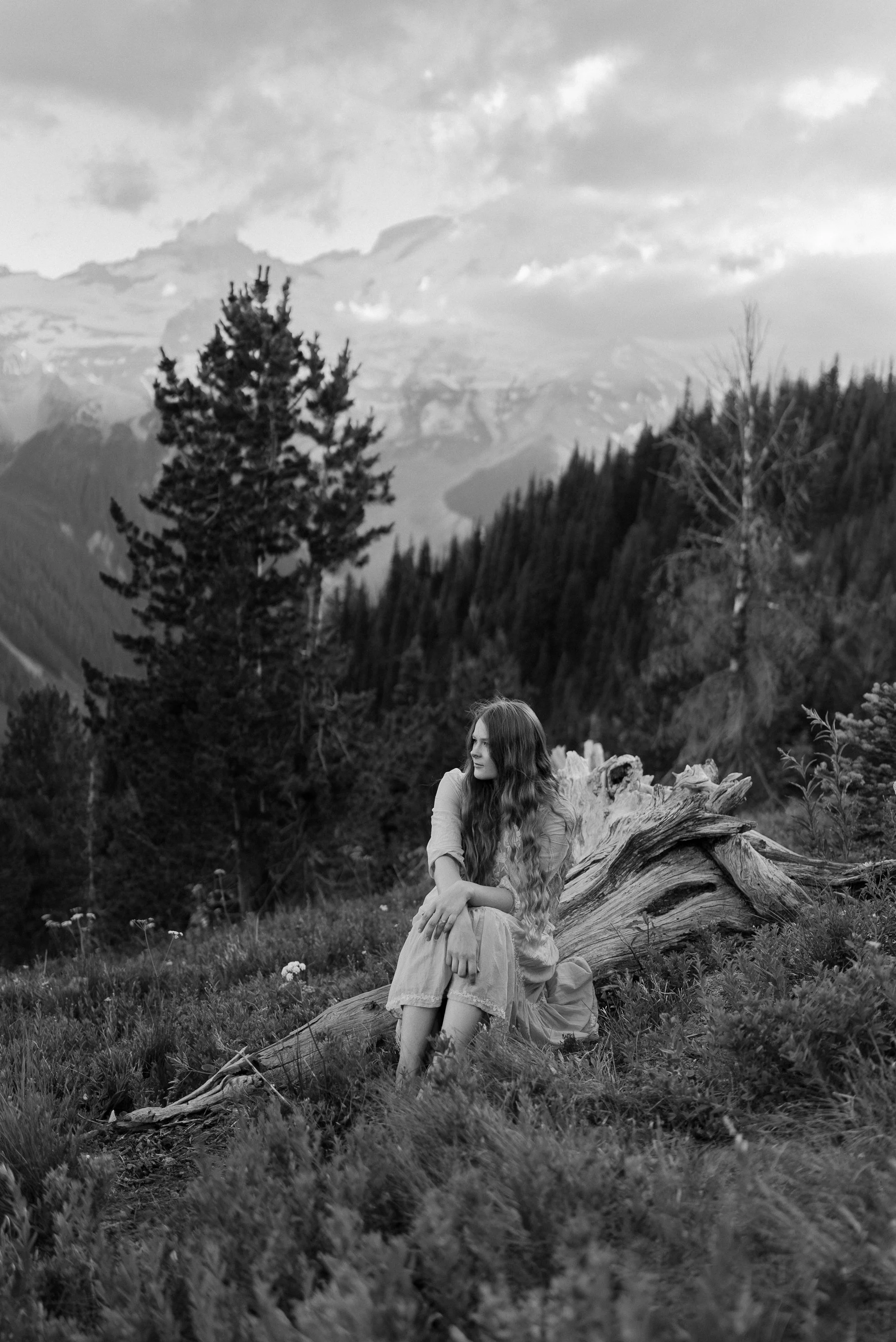 A woman with long hair sitting on a fallen log in a mountain landscape with trees and mountains in the background, in black and white.