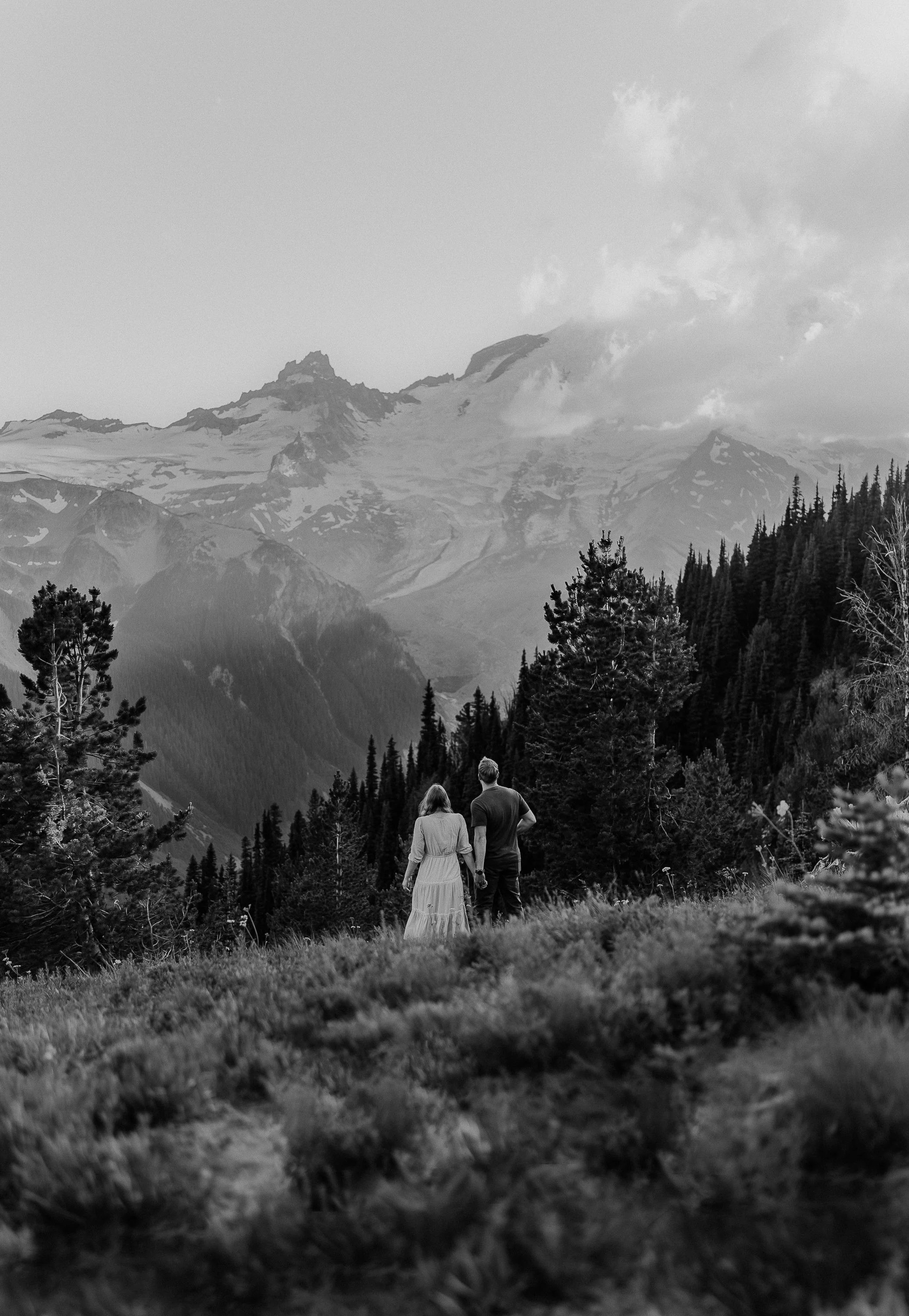 A couple holding hands walking through a grassy field near a forest with mountains in the background, black and white photo.