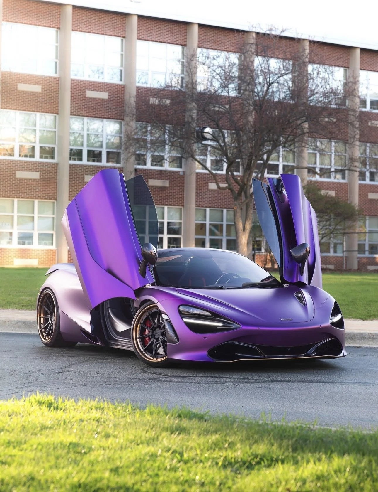 A purple sports car with gullwing doors open parked on the street in front of a brick building with large windows and a tree.