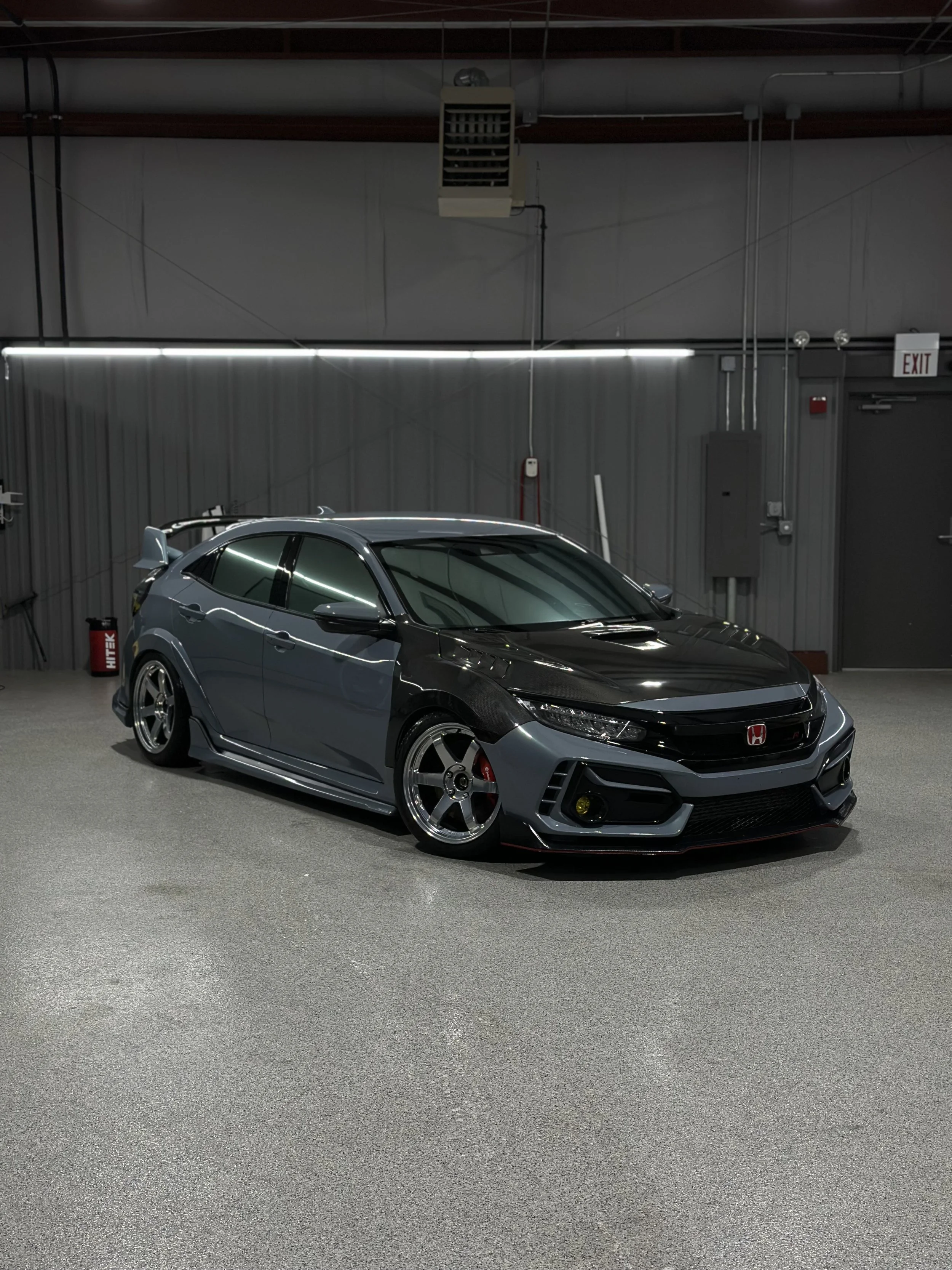 Close-up of a black off-road vehicle with large tires and black and silver rims inside a garage, with a ladder and American flag visible in the background.