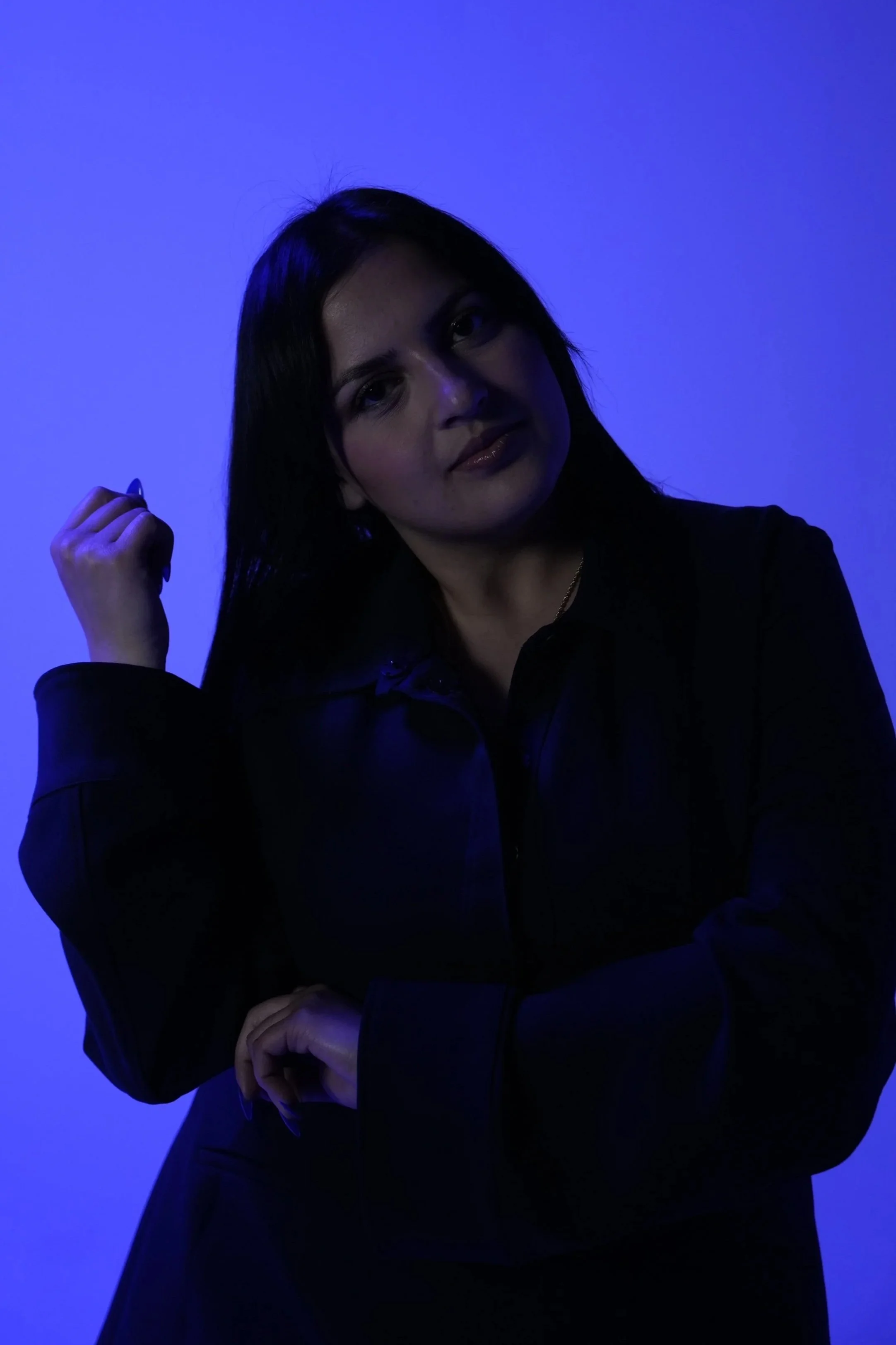 Une femme avec des cheveux noirs, portant une veste noire, posant sous un éclairage bleu, regardant l'appareil photo.