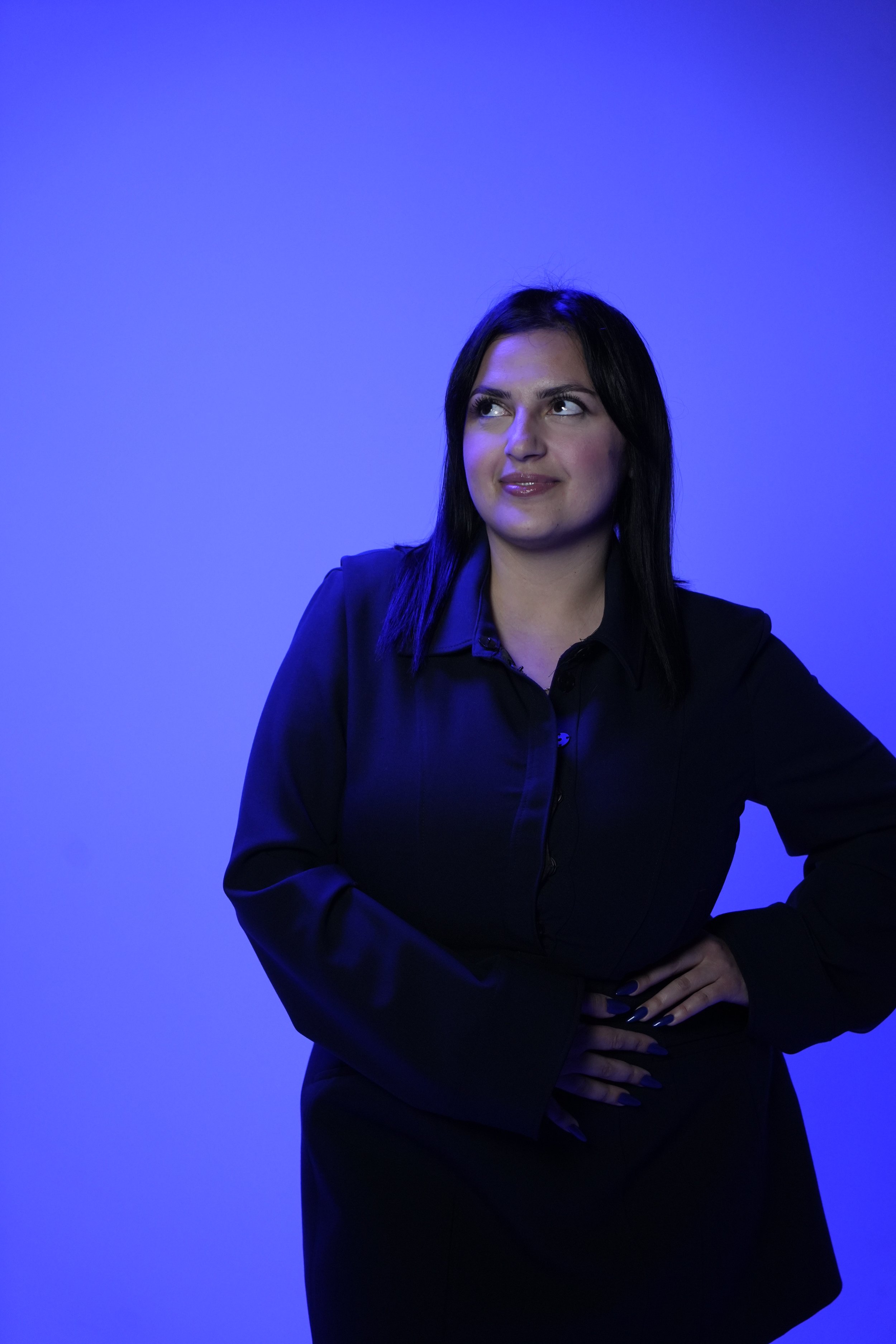 Une femme avec des cheveux noirs, portant une chemise noire, pose devant un fond bleu.
