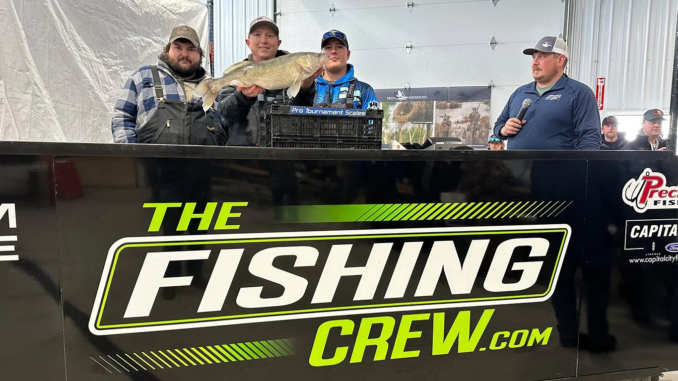 Four men standing behind a black table with the logo 'The Fishing Crew' and holding a large fish. One man in the center is holding the fish with both hands. They are all wearing casual outdoor clothing and caps, and one man on the right is speaking into a microphone.