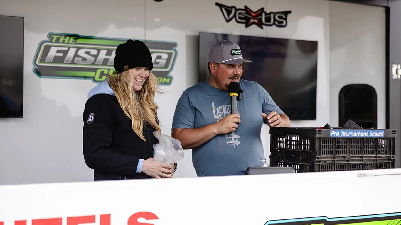 Two people speaking at a fishing competition, one woman holding a plastic bag and a man speaking into a microphone. Behind them are banners for 'The Fishing Crew' and 'Vexus'.