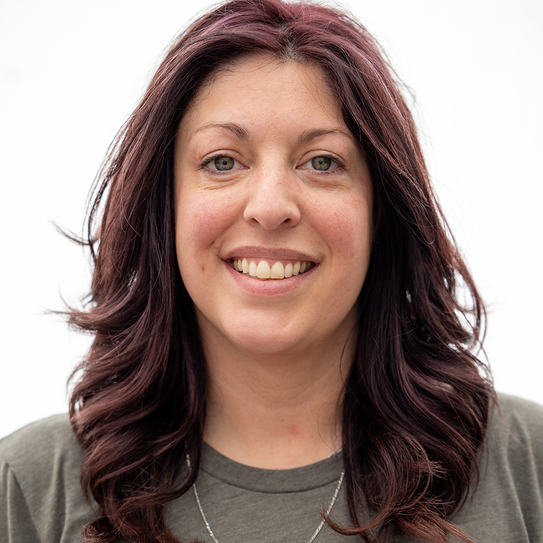 Close-up of a young woman with long, curled dark red hair smiling, wearing a gray top and a silver necklace, against a plain white background.