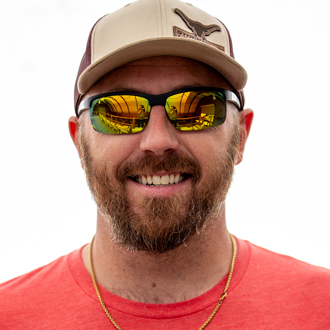 A smiling man with a beard wearing a tan and maroon baseball cap with a bird logo, reflective sunglasses, and a red shirt. The reflection in his sunglasses shows a person taking a photo indoors.
