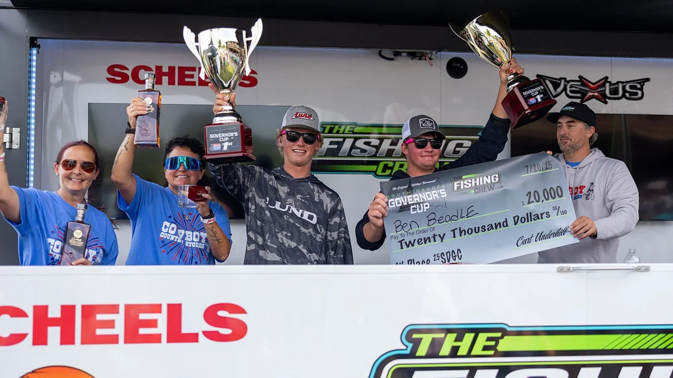 A group of five people on a winners' podium celebrating victory, holding trophies and a large check, with a banner in the background that reads 'The Fishing Crew' and 'Governor's Cup.'