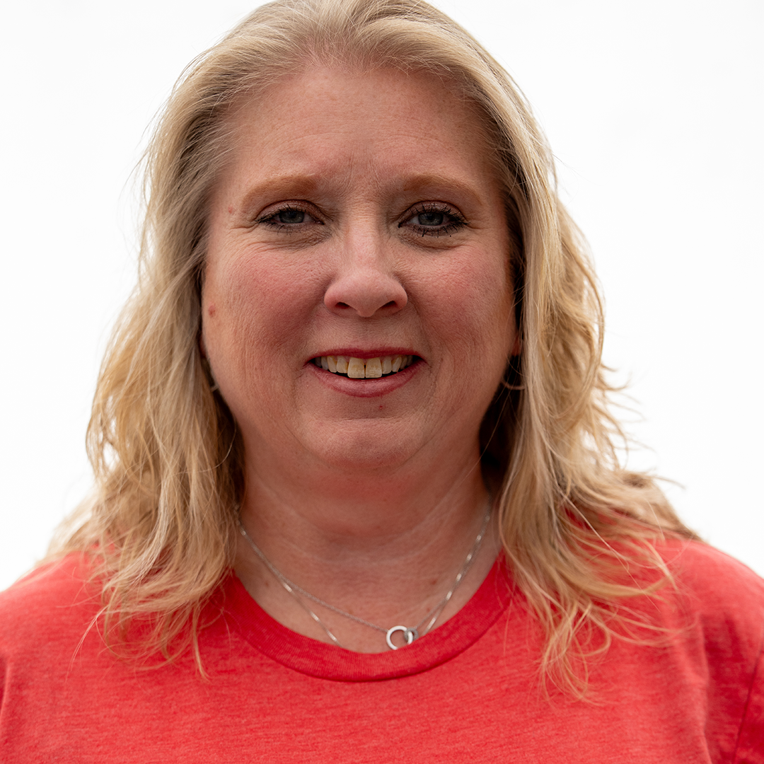 A woman with blonde, wavy hair wearing a red shirt and silver necklaces, smiling against a plain white background.