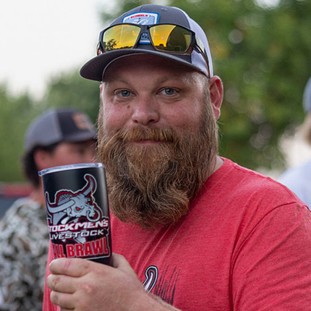 A man with a red beard and mustache wearing a red t-shirt, a cap with sunglasses on top, holding a cup with a Texas Longhorns logo, smiling outdoors with blurred trees in the background.