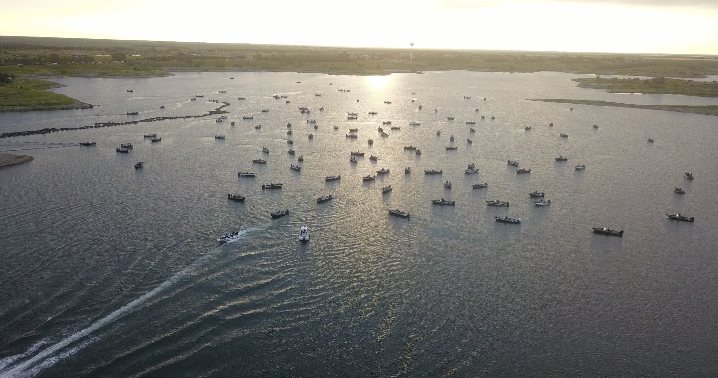 Aerial view of a large body of water with many boats floating and moving, with land visible in the background and a sunset reflecting on the water.