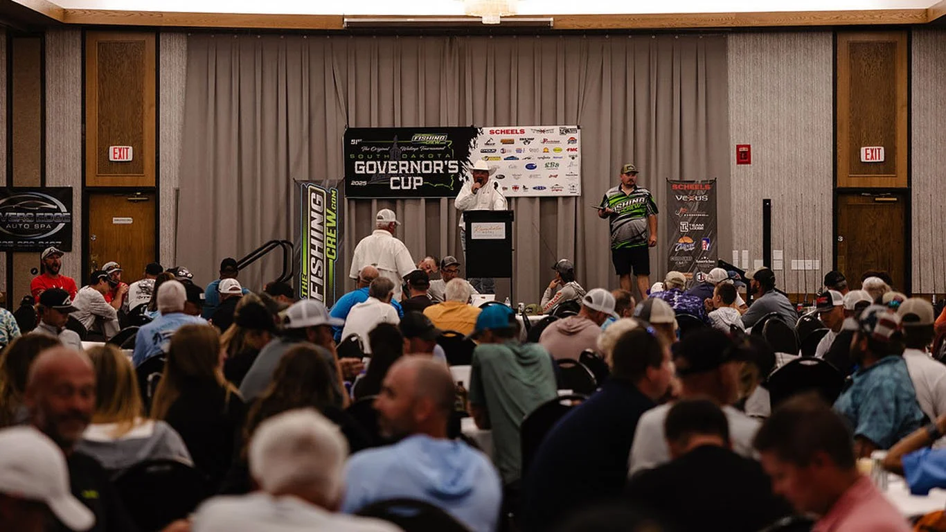 A large crowd attending an event in a conference room, with a stage at the front. On the stage, a person is speaking at a podium, and another person stands nearby. The backdrop includes banners with text, including "Governor's Cup," and various logos. The audience is seated at round tables, and the room has wood-paneled walls.