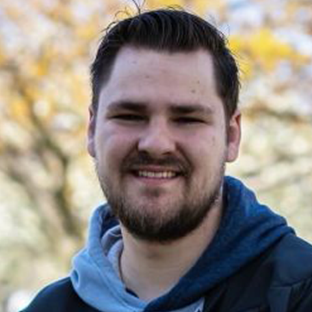 A smiling man with dark hair and a beard outdoors with autumn trees in the background.