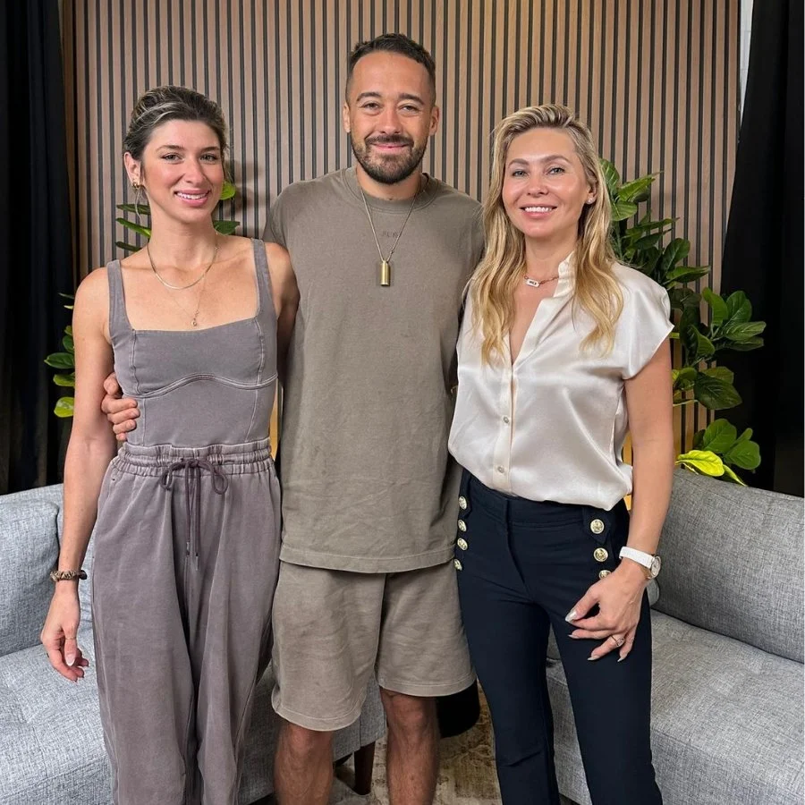 Three people smiling and standing close together in front of a wooden panel background, with green plants on either side.
