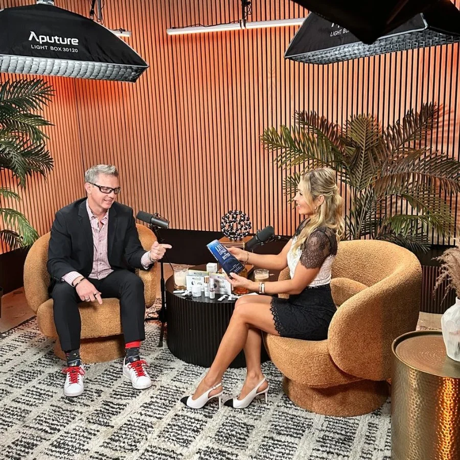 Two people having a conversation in a podcast studio with orange textured walls, green plants, and professional lighting.