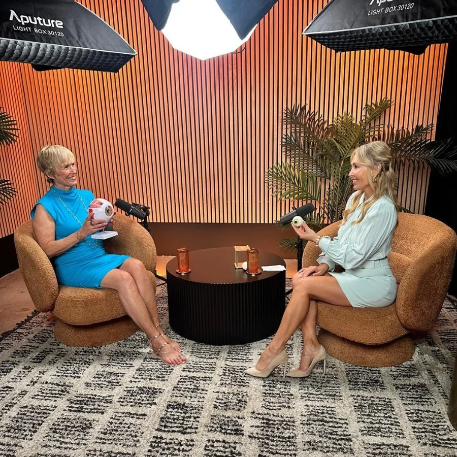 Two women sitting in a studio setting with professional lighting, microphones, and coffee table, appearing to be in an interview or conversation. The woman on the left is wearing a blue dress, holding a prop and smiling, while the woman on the right