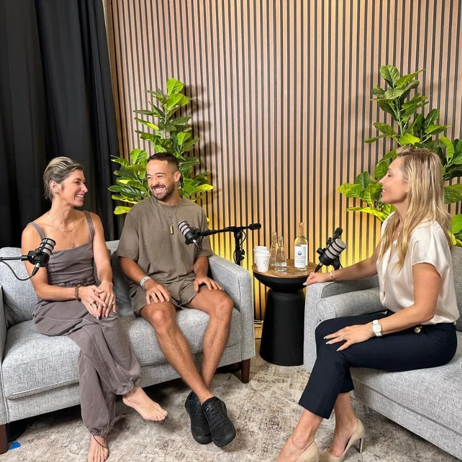 Three people having a conversation in a podcast studio with microphones, water bottles, and drinks on a small table between them, surrounded by green plants and a wooden panel wall in the background.