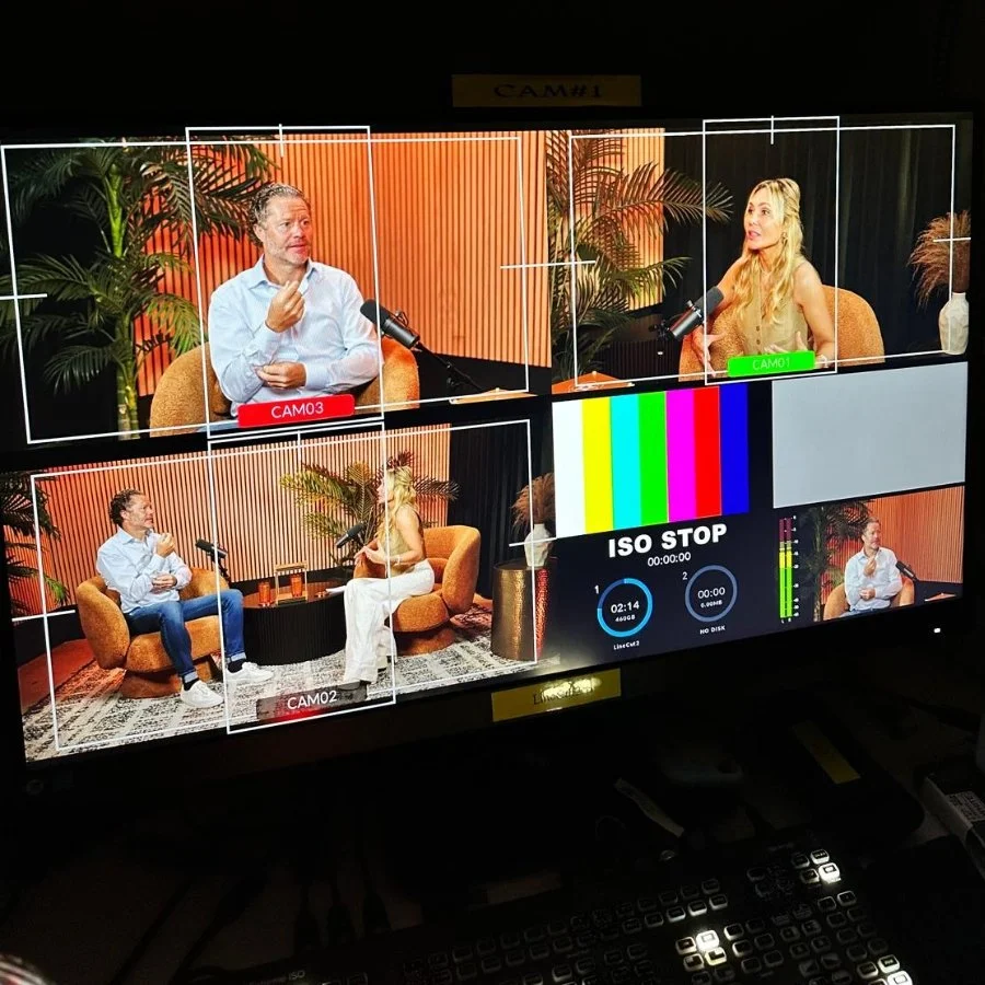 A behind-the-scenes view of a TV interview or podcast recording studio with two people seated on orange chairs, surrounded by large green plants and warm lighting. The monitor shows multiple camera angles, a color test pattern, and technical displays