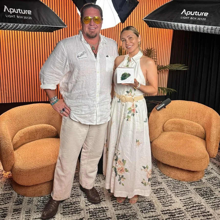 A man and woman standing together in a photography studio with professional lighting equipment. The man is wearing a white shirt, beige pants, and sunglasses, and the woman is wearing a floral dress and holding a box with greenery on it. They are smi