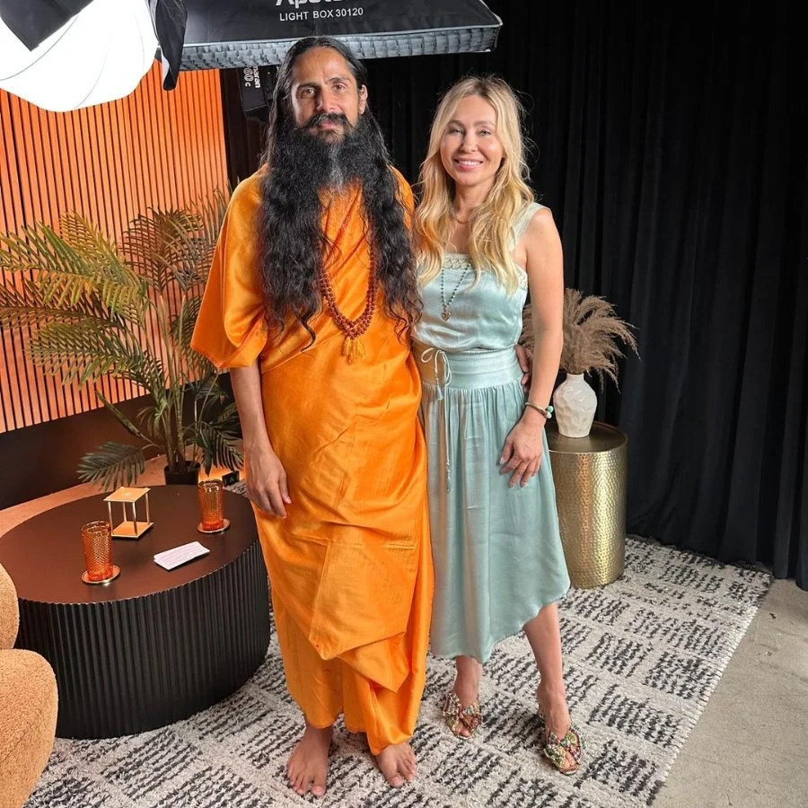 A man in orange traditional attire with a long beard and long hair next to a woman in a light blue dress with blonde hair, standing together and smiling inside a room with decorative plants, a black curtain, and modern furniture.