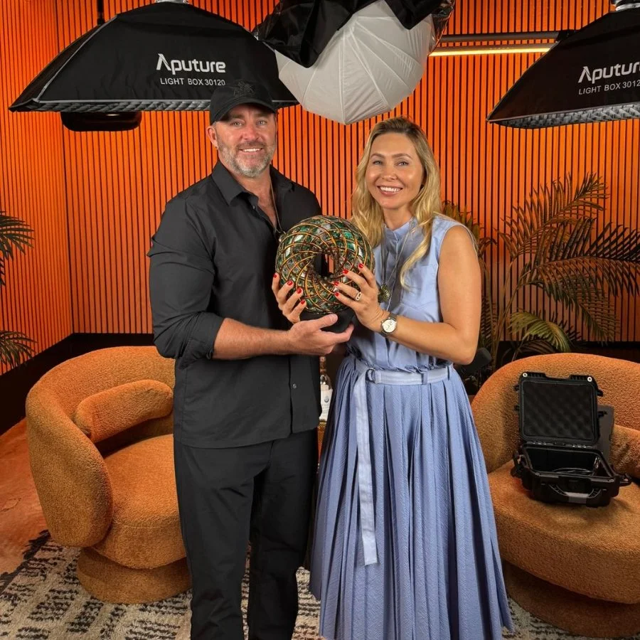A man and woman stand together in a photography studio, holding a colorful spherical object. The man has a beard, is wearing a black shirt and a black cap, and the woman has long blonde hair, is dressed in a light blue dress, and is wearing a watch.