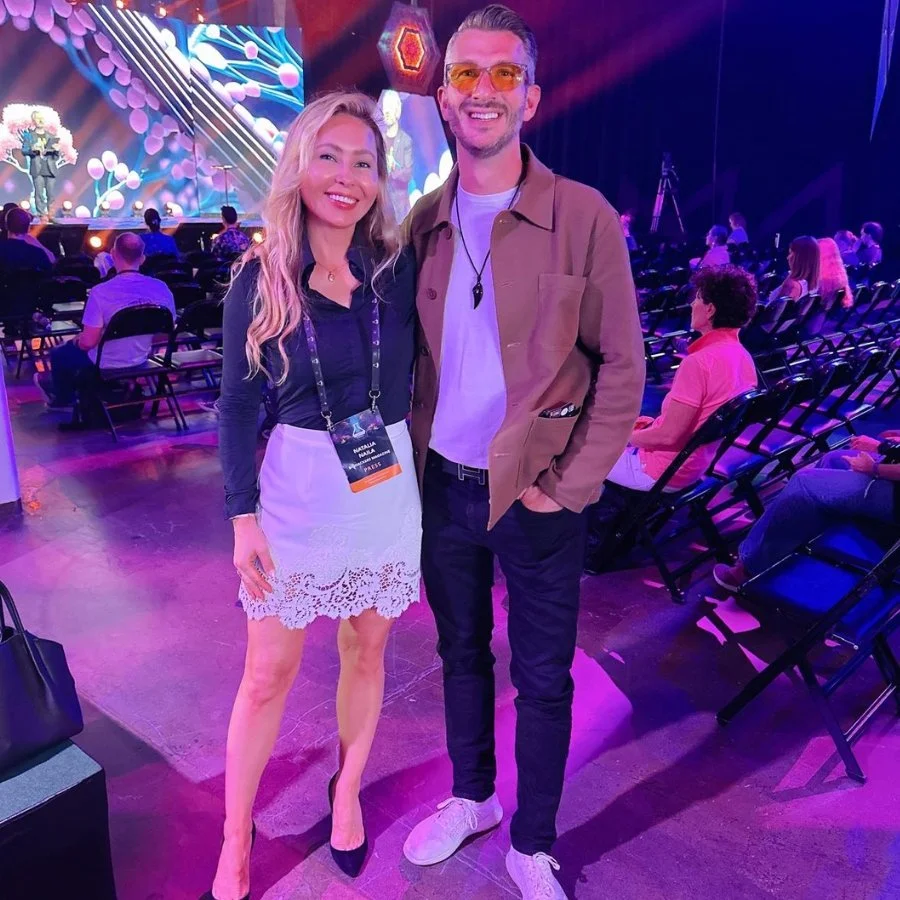A woman and a man smiling for a photo at an indoor event with stage lighting and a seated audience.