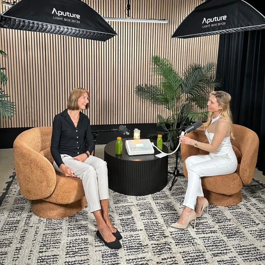 Two women engaged in an interview or conversation in a recording studio, sitting on tan armchairs with a black table between them, microphones, and green bottles on the table, with light wood-paneled walls, plants, and professional studio lighting ov