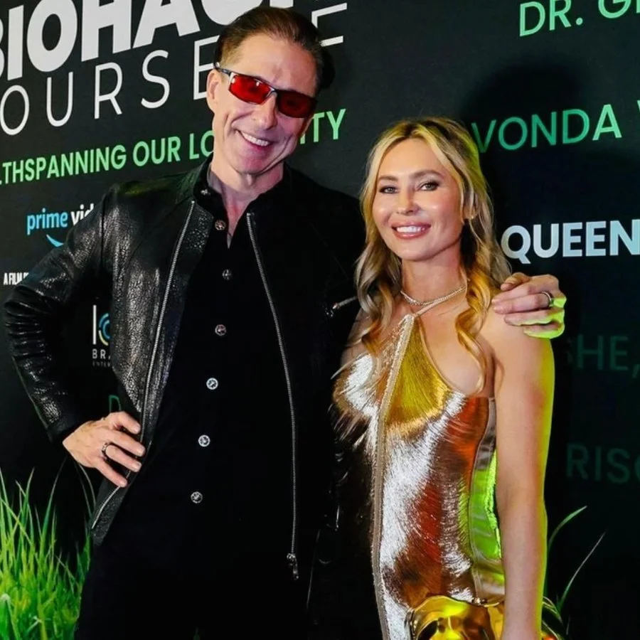 Two people, a man and a woman, pose together at an event, standing in front of a black background with green text and logos. The man is wearing a black leather jacket and red sunglasses, smiling with his arm around the woman's shoulder. The woman is