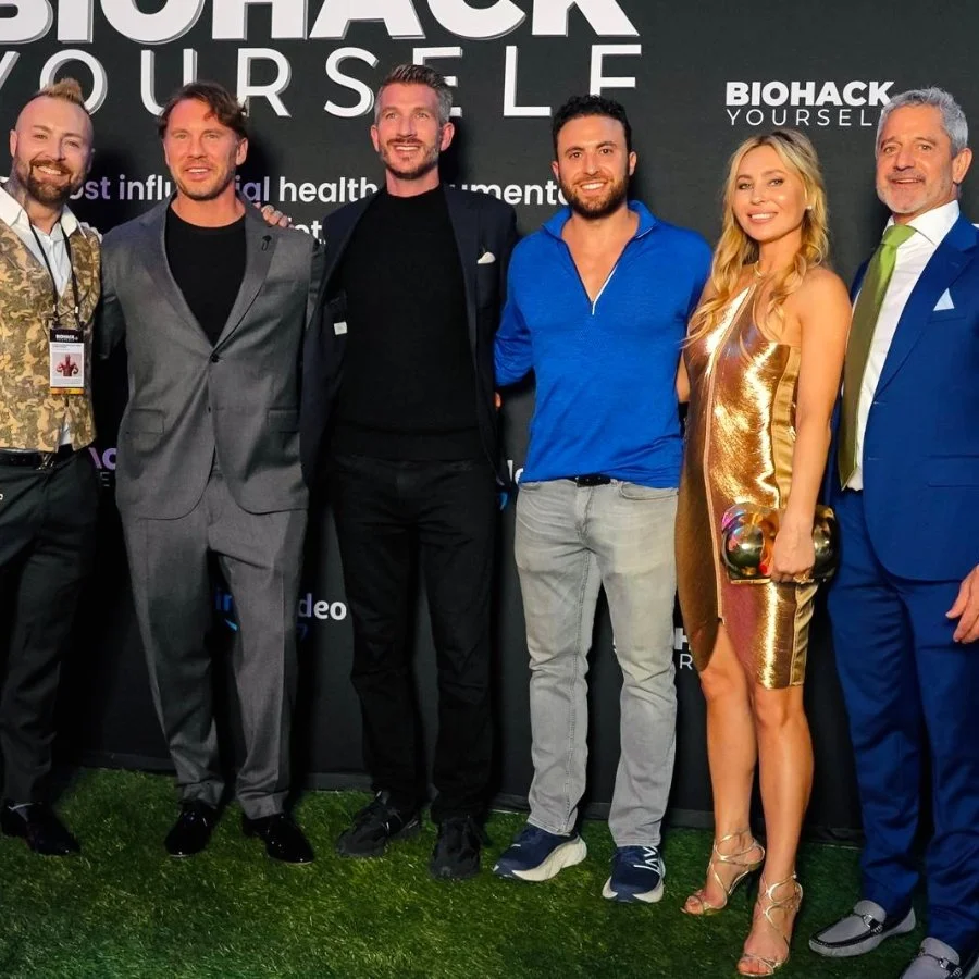 A group of six people posing for a photo at a Biohack Yourself event, dressed in formal and semi-formal attire, standing in front of a black backdrop with white text.