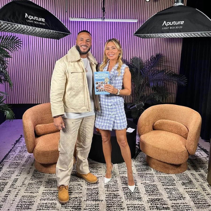 Two people smiling and posing for a photo in a modern indoor setting with purple and black striped walls, flanked by large studio lights and potted plants. One person is holding a book titled 'Modern Wellness'.