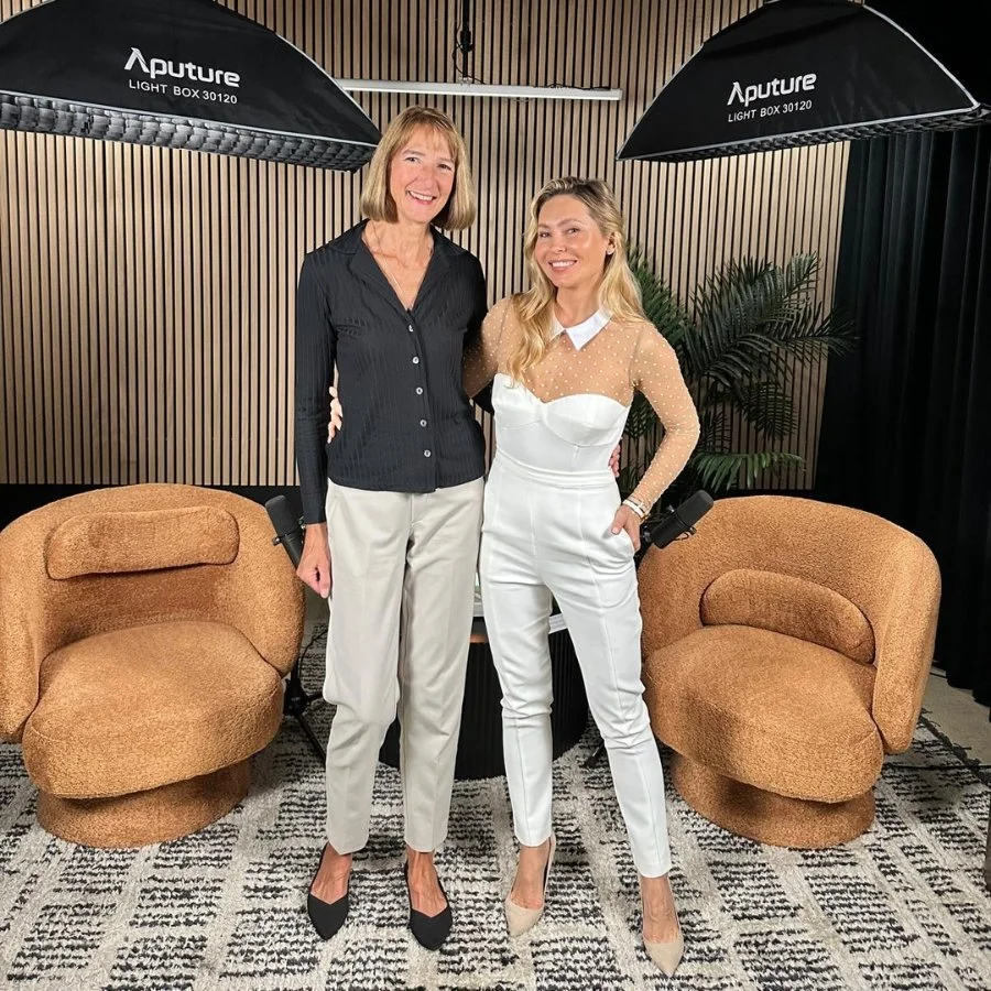 Two women standing side by side in a studio, smiling at the camera. They are surrounded by studio lighting equipment labeled 'Aputure' and a background with vertical wooden panels. One woman is dressed in a black blouse and beige pants, and the other