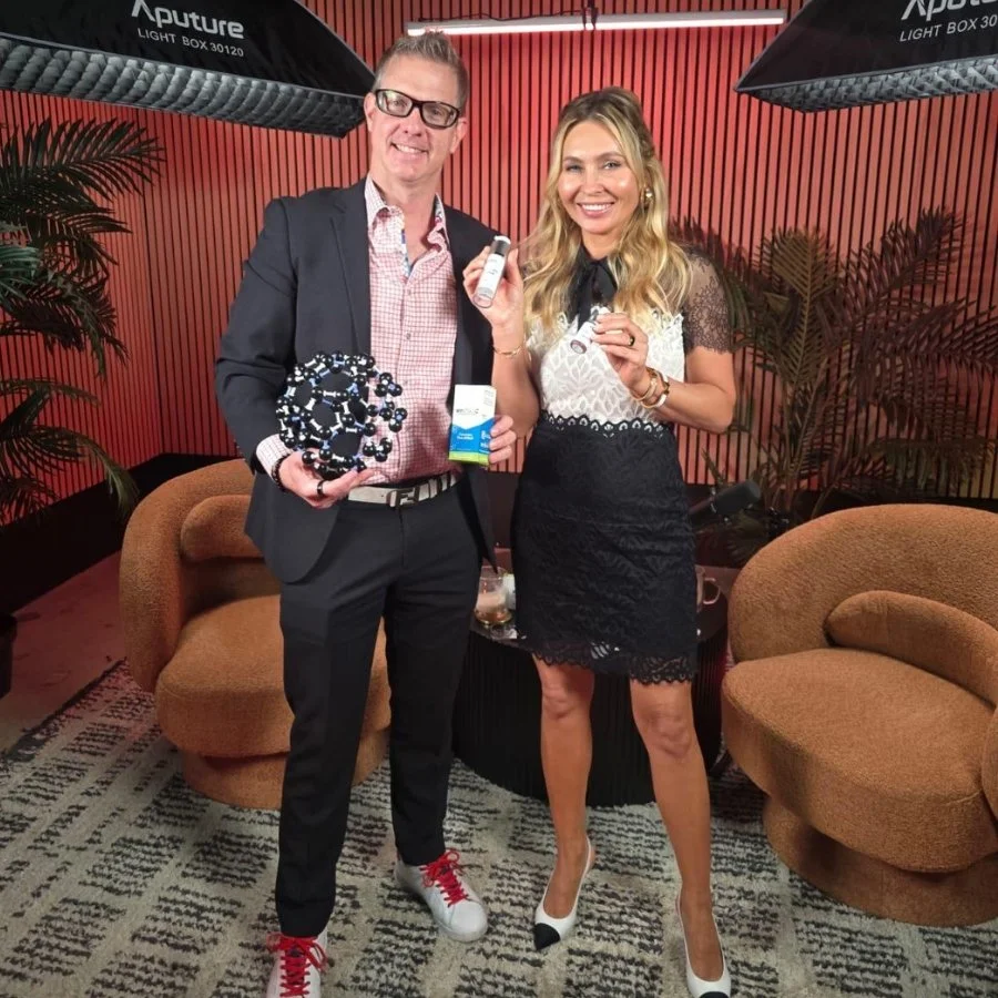 A man and woman smiling and posing together indoors. The man holds a molecular model and a small box, dressed in a dark suit with a checked shirt and red sneakers. The woman wears a black and white lace dress with high heels. The background features