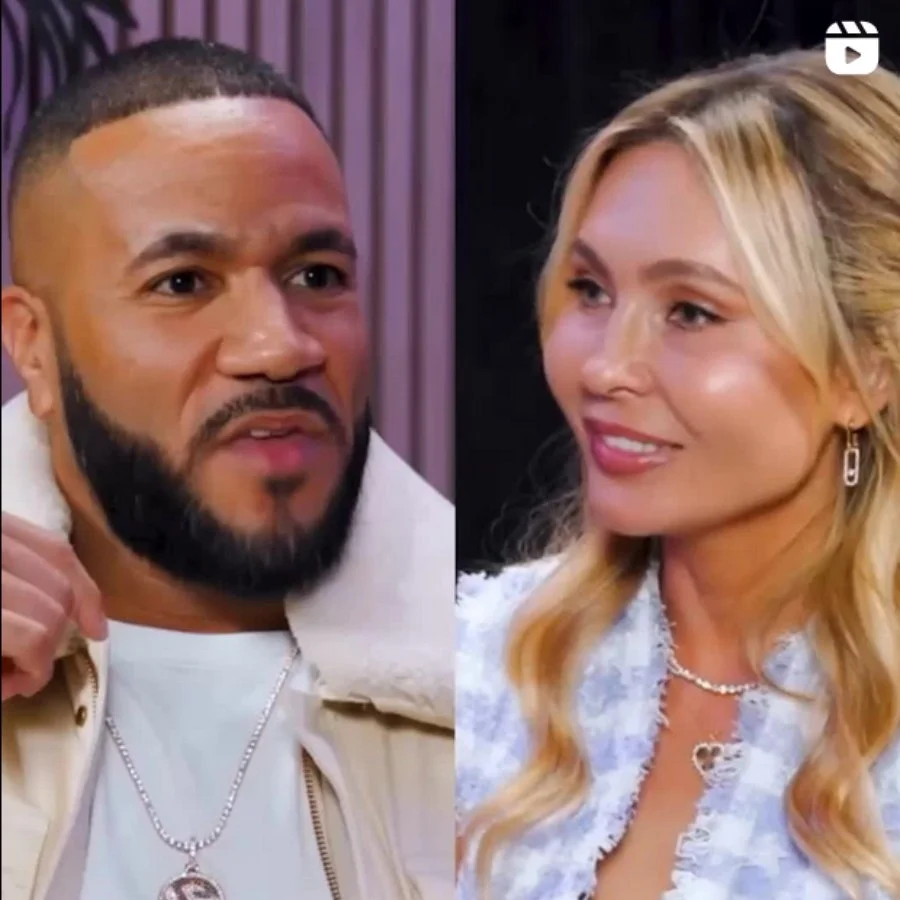 Close-up of a man and woman facing each other, engaging in conversation. The man has a beard, short hair, and is wearing a light-colored jacket with a chain necklace. The woman has long blonde hair, earrings, a necklace, and is wearing a light blue o