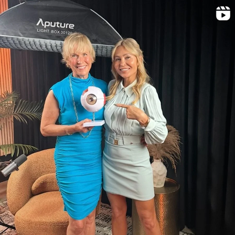 Two women smiling and posing indoors; one holding a decorative object resembling an eye, while the other points at it; background includes a black curtain, a potted plant, and a conversation area with a beige chair and a side table.