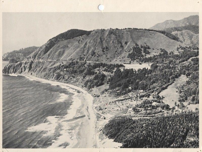 A black and white aerial photograph showing a coastal scene with waves hitting the shoreline, a beach, and steep hills covered in trees and grass. There are a few buildings and a winding road visible along the coast.