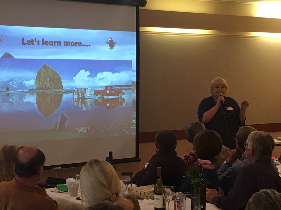 A woman giving a presentation in front of an audience in a conference room. A slide on a projector screen shows an island, a red truck, and the text "Let's learn more..."