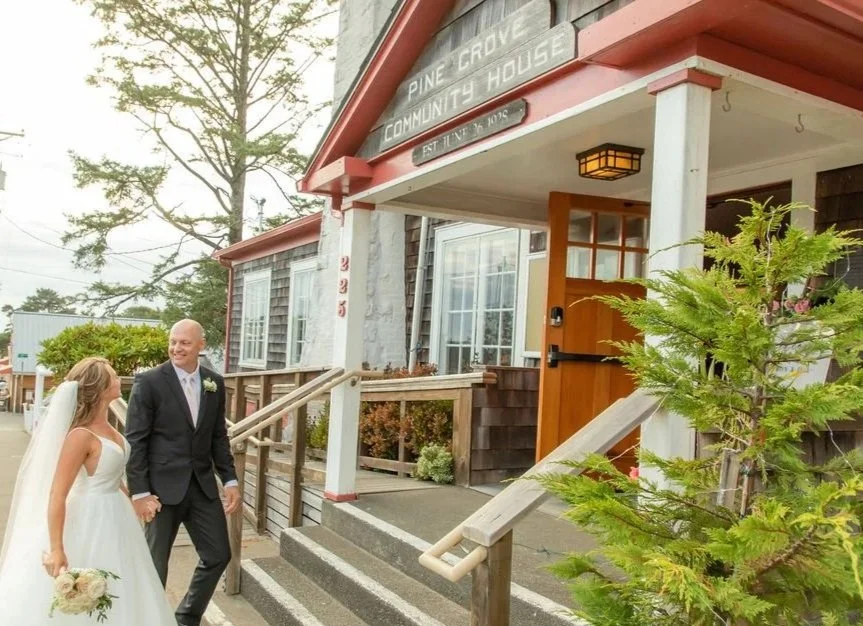 A newlywed couple holding hands and smiling outside The Pine Grove Community House, a rustic venue with a wooden entrance, greenery, and large windows, during daytime.